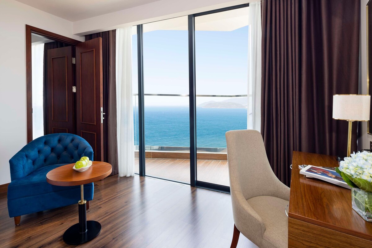 A hotel room with a large sliding glass door leading to a balcony with a sea view. There is a blue armchair, a beige armchair, and a small round table with a bowl of fruit.