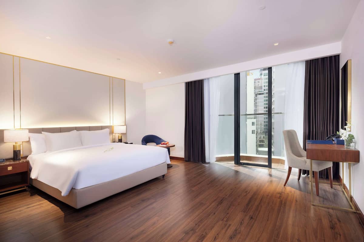 A modern hotel room with a large bed, a desk, and a view of the city through large windows.