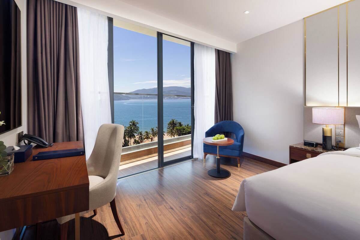 A hotel room with a large window, a desk, a chair, a bed, and a small table with a lamp.
