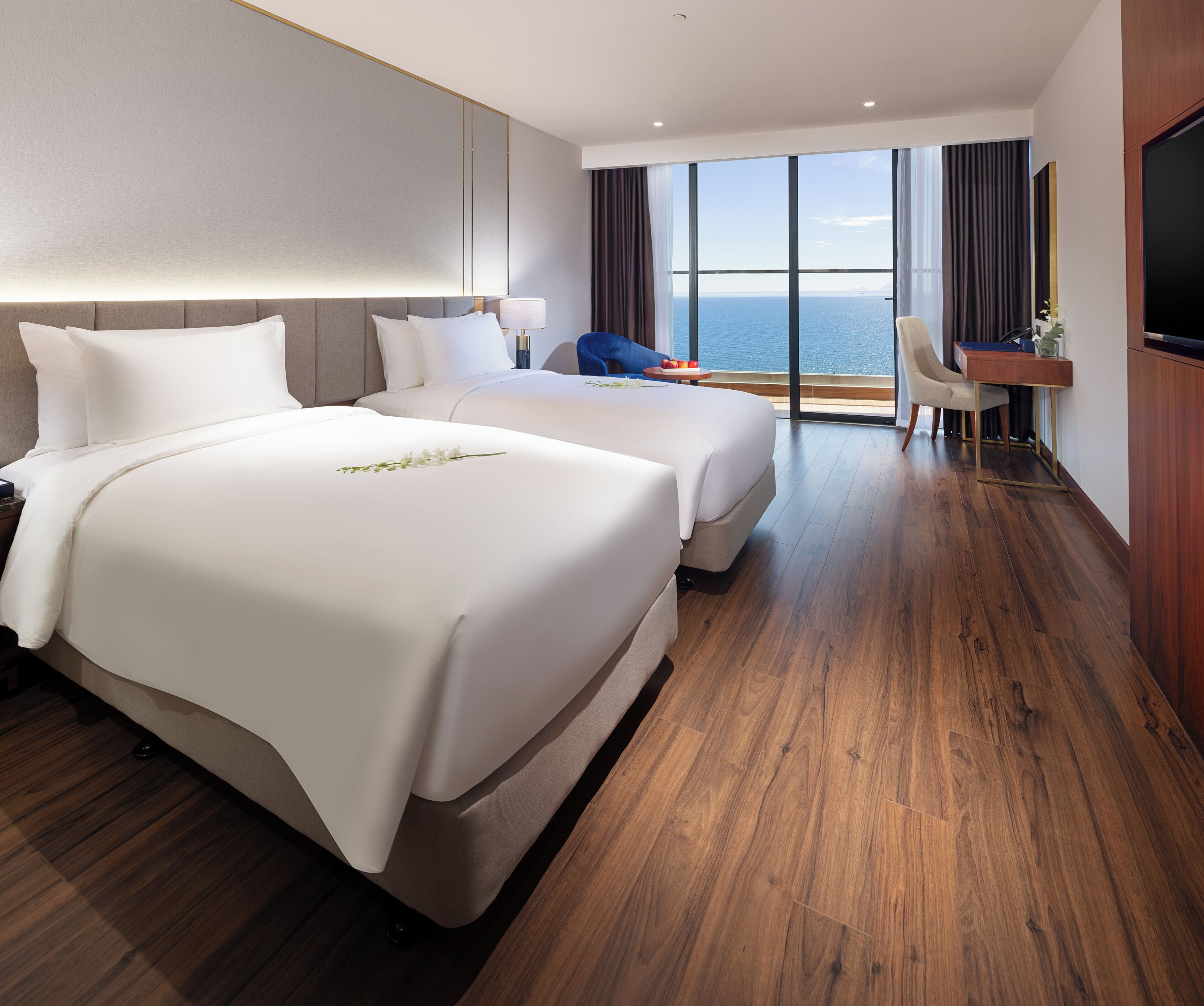 A hotel room with two beds, a desk, a chair, and a large window with a view of the sea.