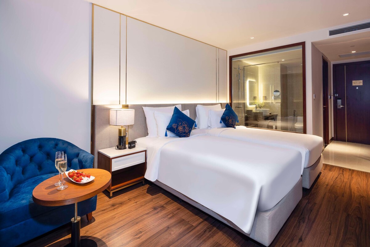A hotel room with a large bed, a bedside table with a lamp, a blue armchair, and a small round table with a glass of water and a plate of strawberries.