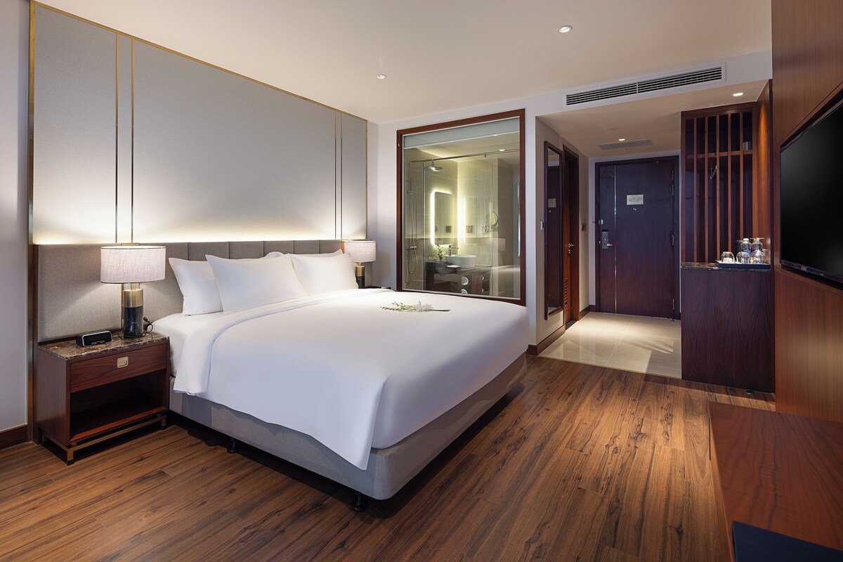A hotel room with a large bed, bedside tables, a TV, and a bathroom visible through a glass door.