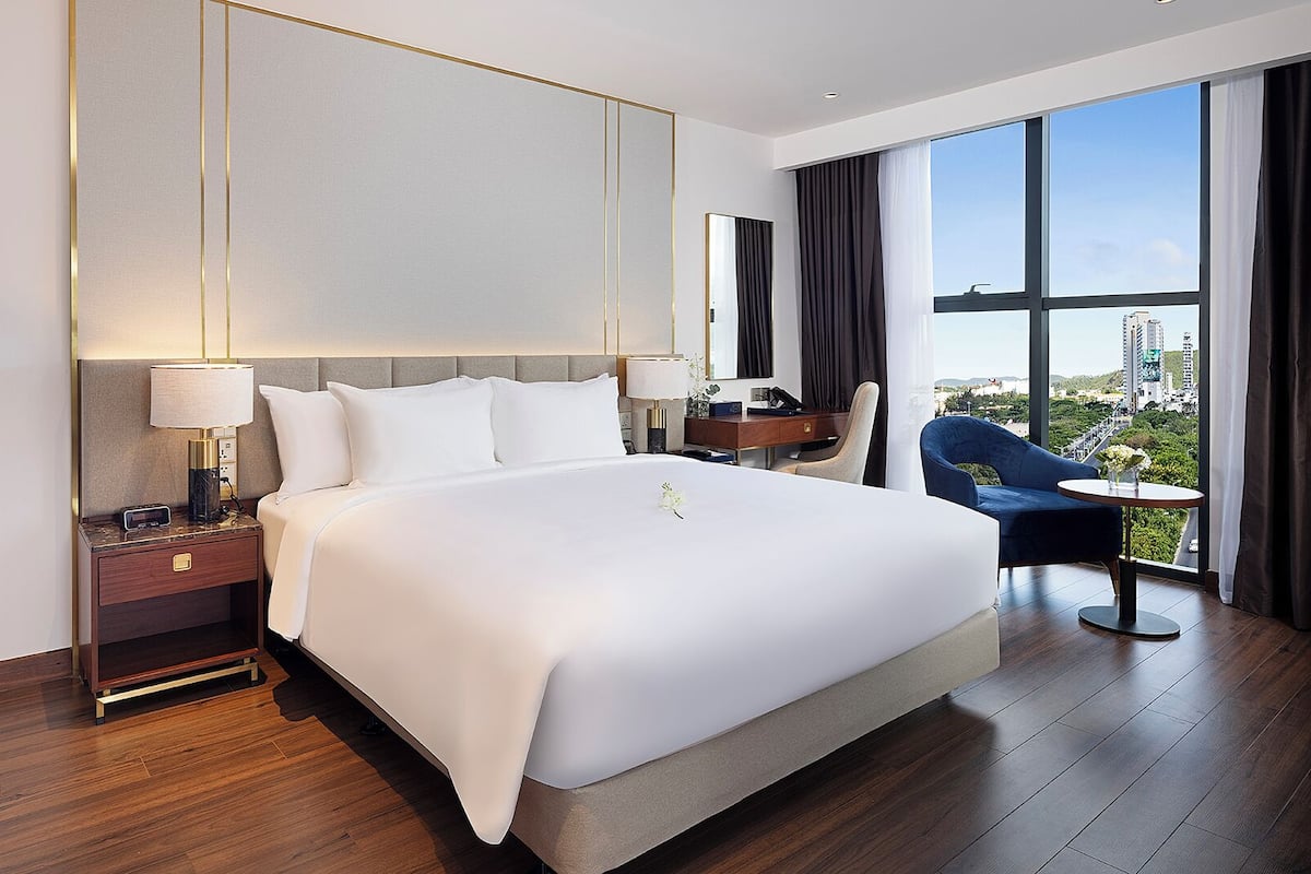 A modern hotel room with a large bed, a desk, a chair, and a view of the city.