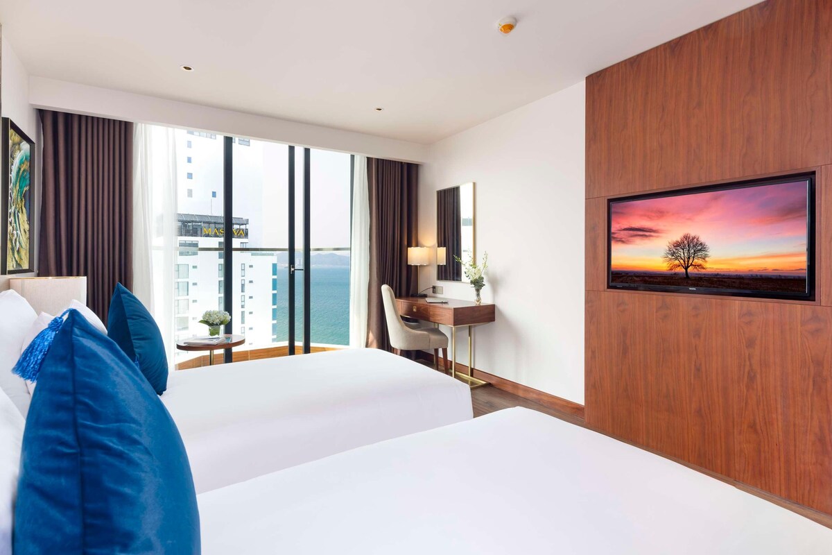 A hotel room with a large bed, a desk with a chair, a TV on the wall, and a view of the city.