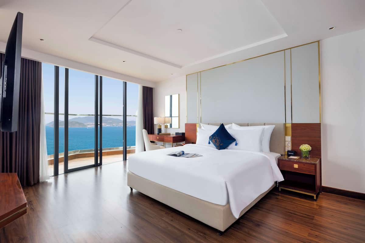 A hotel room with a large bed, a desk, a chair, a TV, and a view of the sea.