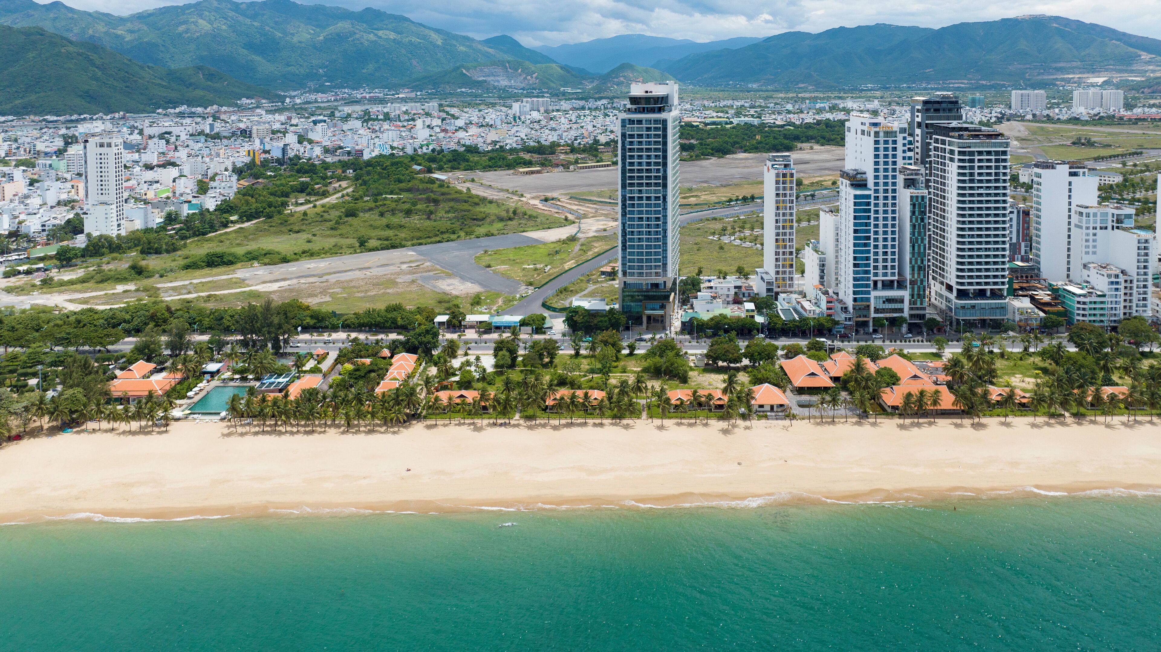 A coastal cityscape with high-rise buildings, a beach, and a mountainous backdrop.