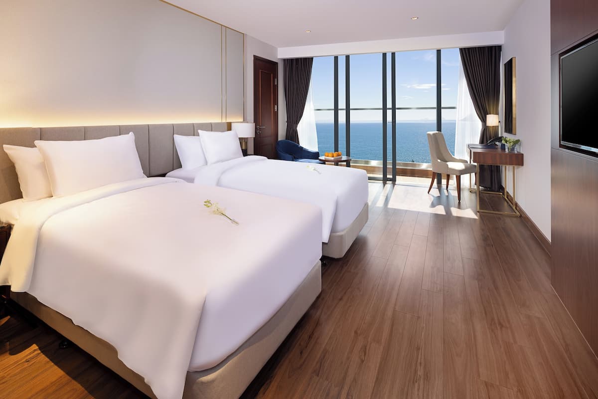 A hotel room with two beds, a desk with a TV, and a view of the sea.