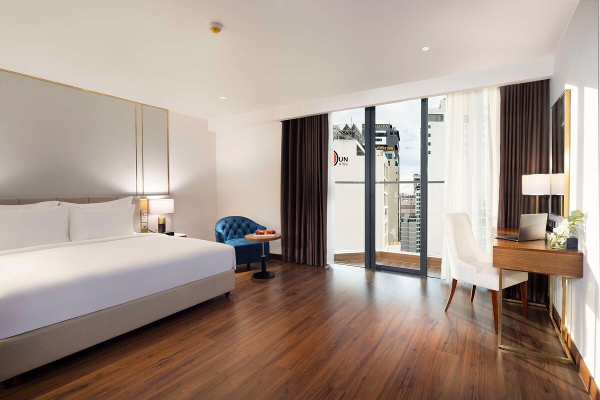 A modern hotel room with a large bed, a desk, and a view of the cityscape through the window.