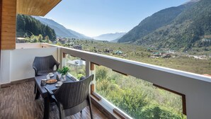 A balcony with wicker furniture, a table set with a teapot, cups, and a plate of food, overlooking a valley with mountains and scattered buildings. - DLS Serenity Resort And Spa (Manali)