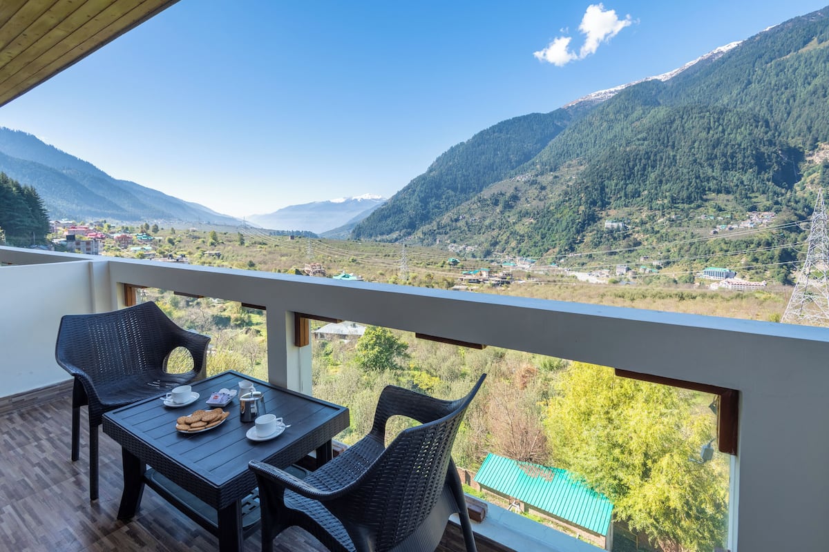 Ein Balkon mit Korbmöbeln, einem Tisch gedeckt mit Tellern und Tassen sowie einem Panoramablick auf die Berge.