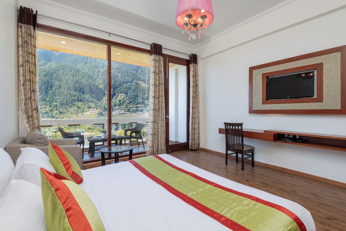 Ein Hotelzimmer mit einem großen Bett, einem an der Wand montierten Fernseher, einem Balkon mit Blick auf einen bewaldeten Berg und einem Essbereich mit Tisch und Stühlen.