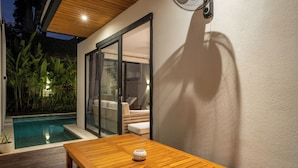 Una mesa de madera con un objeto blanco encima, un ventanal de vidrio que da a una zona de estar y una piscina al fondo.