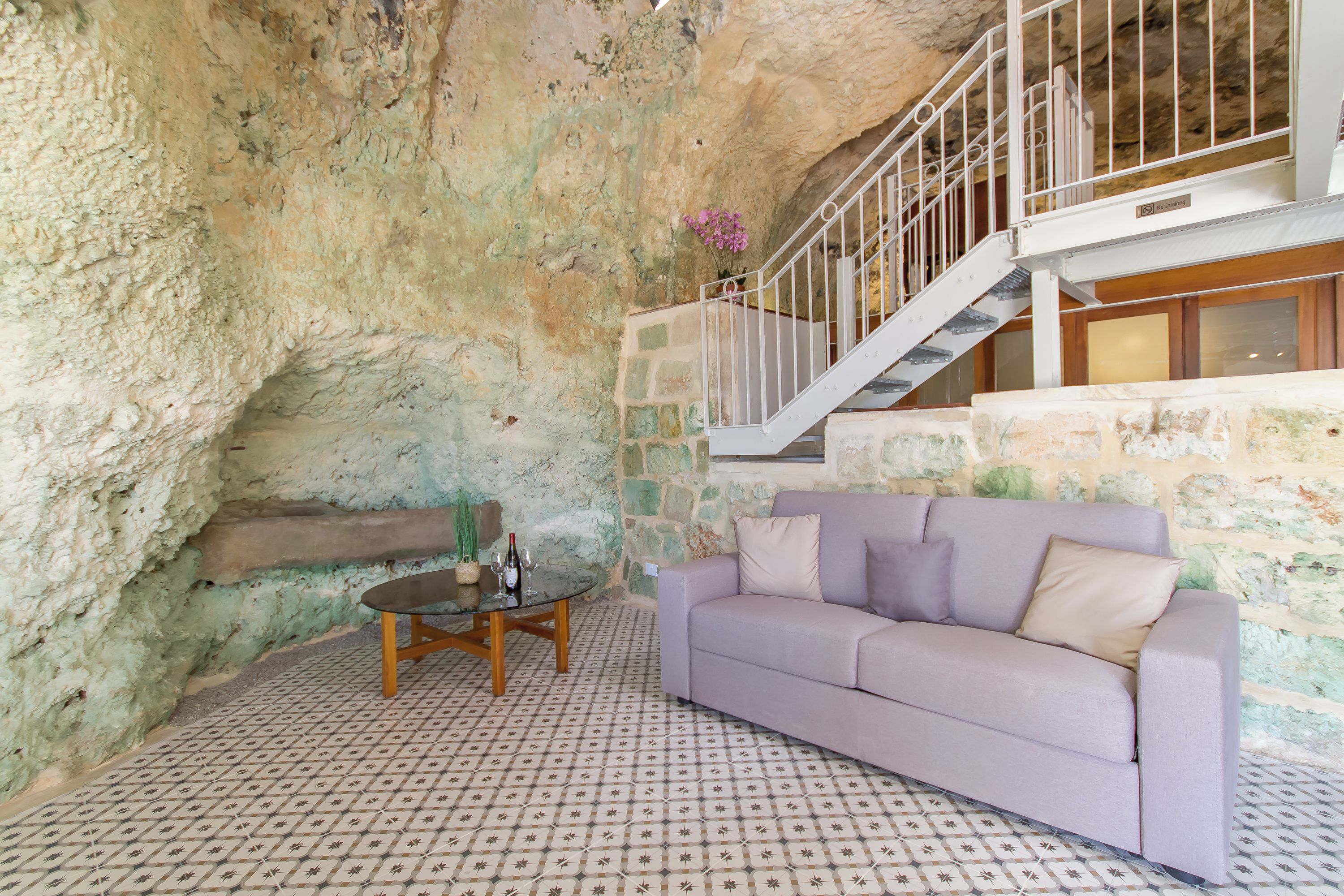 Una habitación con pared de piedra, un sofá morado, una mesita con una botella y una escalera.