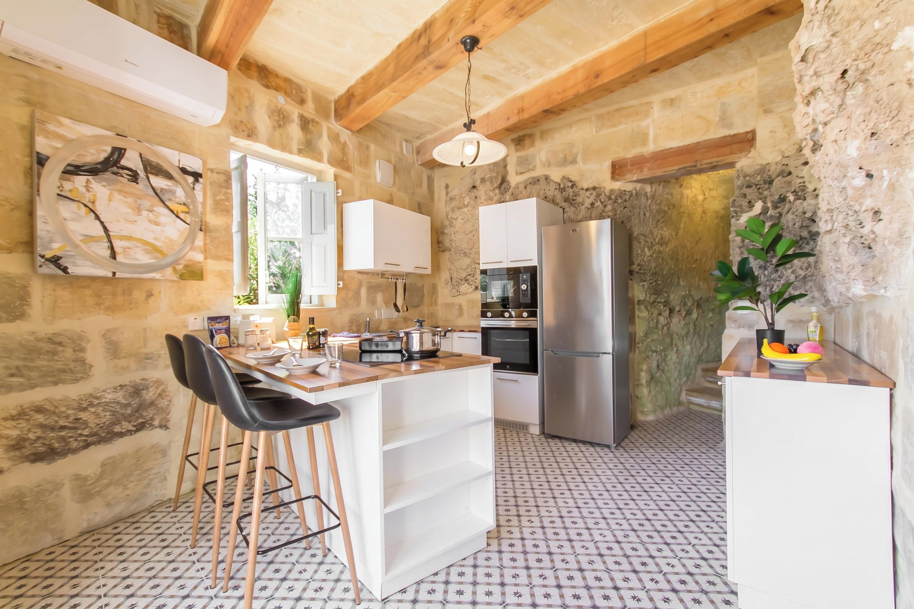 A rustic kitchen with stone walls, a wooden ceiling, a central island with stools, white cabinets, a stainless steel refrigerator, and a patterned floor.