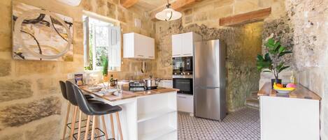 A rustic kitchen with stone walls, a wooden ceiling, a central island with stools, white cabinets, a stainless steel refrigerator, and a patterned floor.