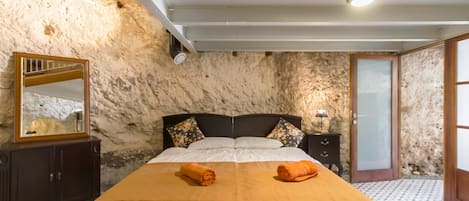 A bedroom with a stone wall, a bed with a white and orange bedspread, a wooden nightstand, a mirror, and a door.