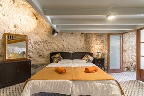 A bedroom with a stone wall, a bed with a white and orange bedspread, a wooden nightstand, a mirror, and a door. - Pleasant Stone House & Jacuzzi St Martin (Mgarr)
