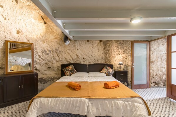 A bedroom with a stone wall, a bed with a white and orange bedspread, a wooden nightstand, a mirror, and a door.