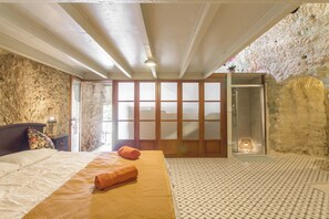 Quarto com cama, mesa de cabeceira, porta de madeira e parede de pedra.