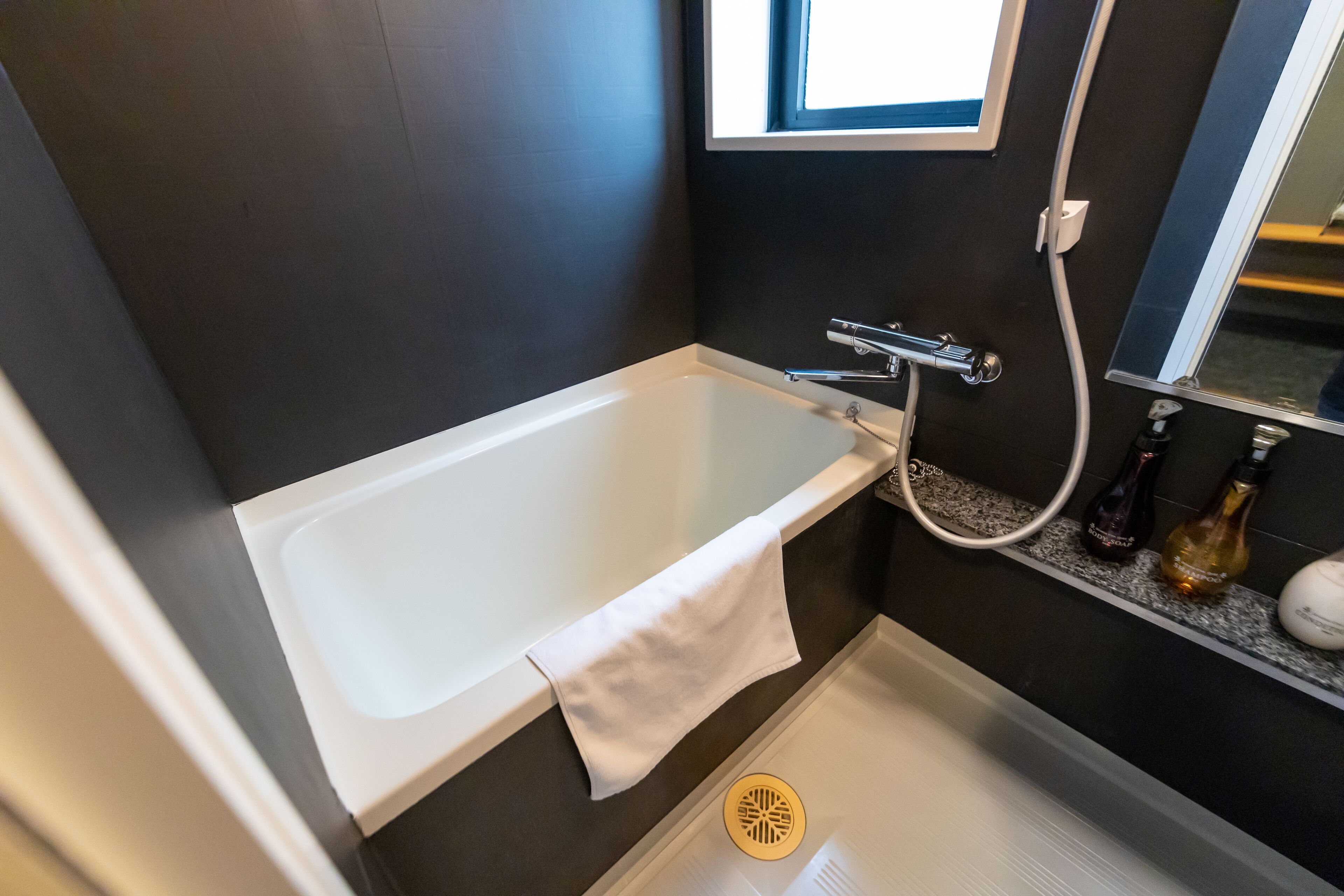 A modern bathroom with a bathtub, a towel, a window, and a shelf with bottles.
