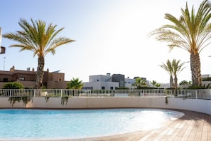 A swimming pool with a wooden deck, surrounded by palm trees and modern buildings in the background.