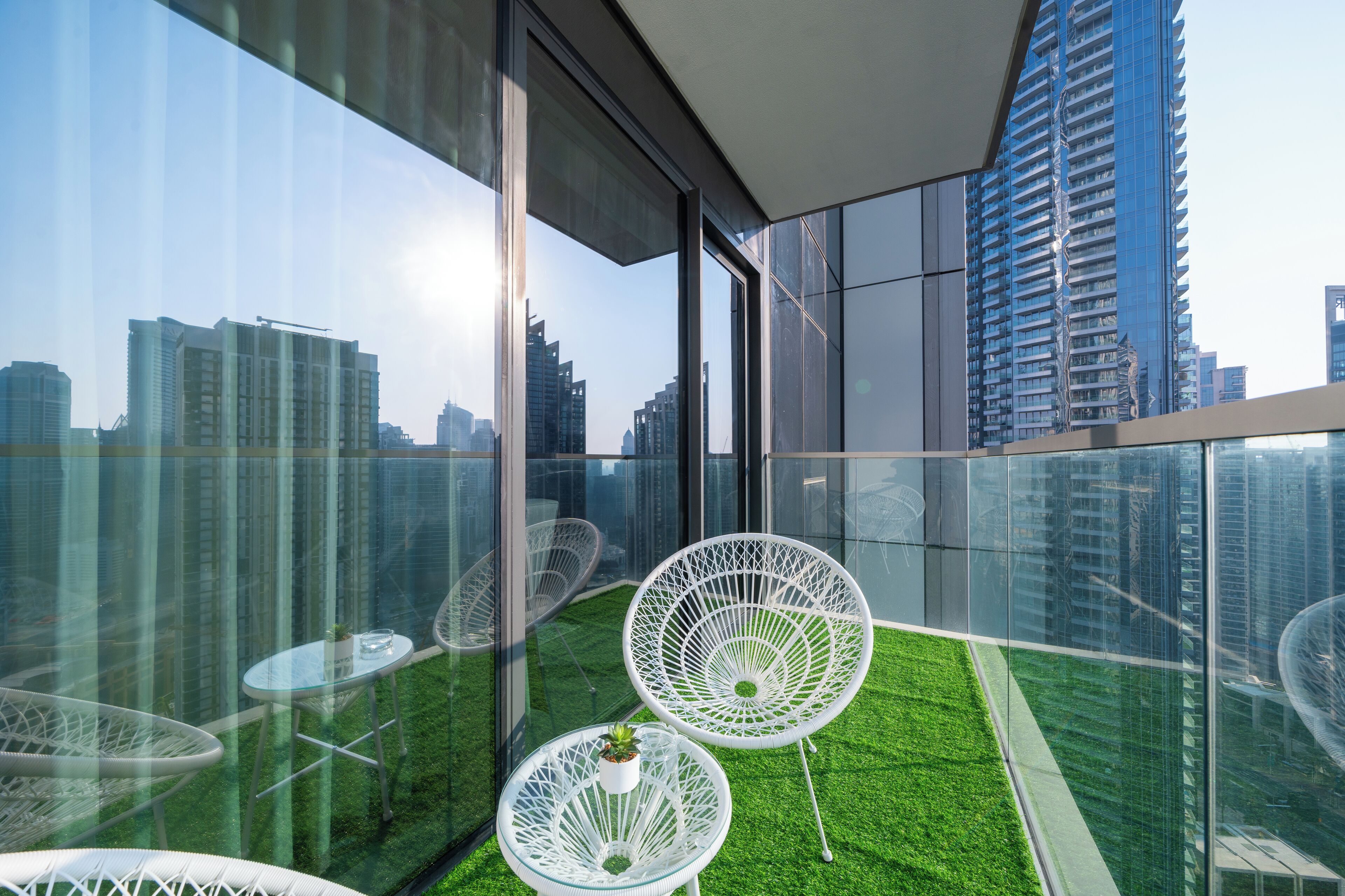Un balcon moderne avec des murs en verre, des chaises blanches et une petite table agrémentée d’une plante.