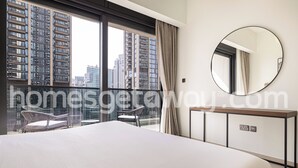 Camera d'albergo con un letto grande, uno specchio tondo, un tavolino e vista su alti palazzi attraverso ampie finestre.