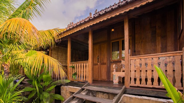 Una casa de madera con un porche, escalones que llevan a la entrada y plantas tropicales alrededor.