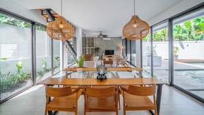 Uma sala de jantar moderna com mesa de vidro e cadeiras de madeira, janelas amplas e escada que leva a um andar superior.