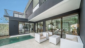 Uma área externa moderna com piscina, móveis de vime branco e portas de vidro que dão acesso a um espaço interno.