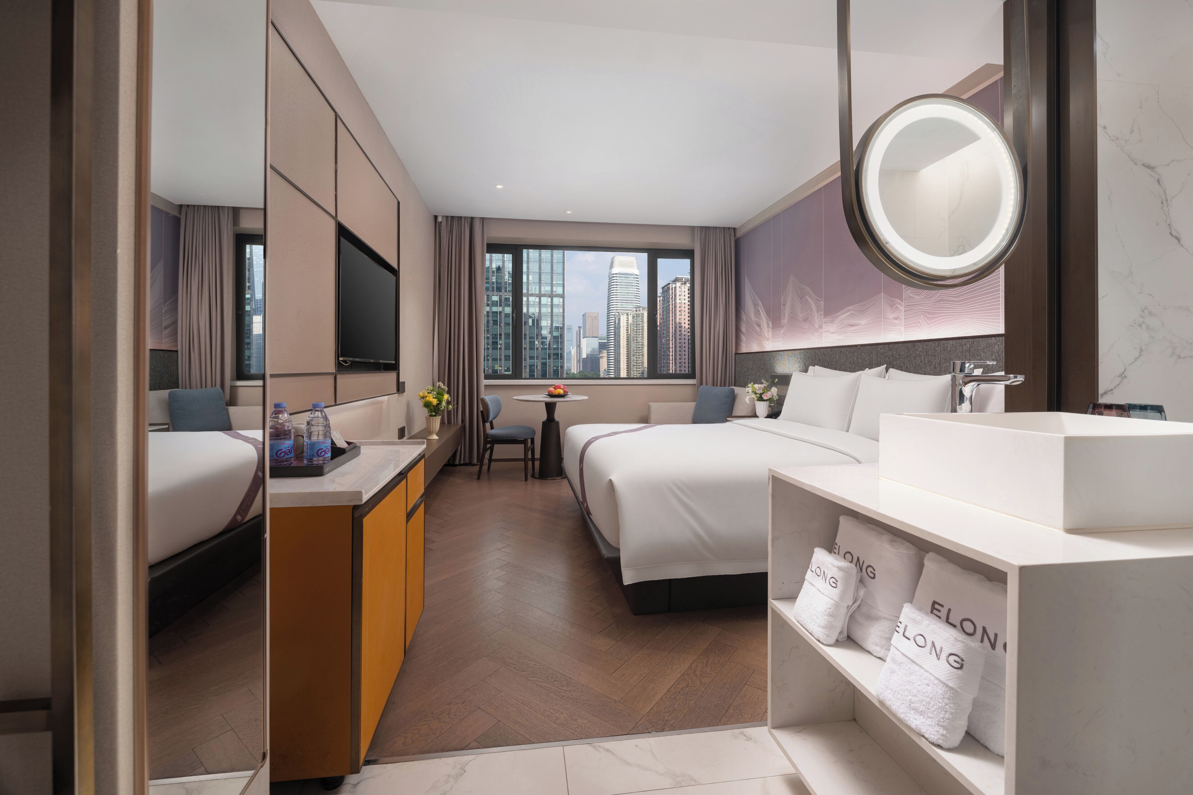 A hotel room with a bed, a desk, a chair, a round mirror, and a bathroom with a sink and towel rack.