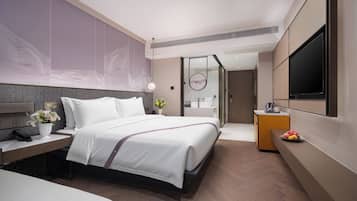 Una habitación de hotel moderna con una cama grande, una mesita de noche, un televisor montado en la pared y un baño visible a través de una puerta de cristal.
