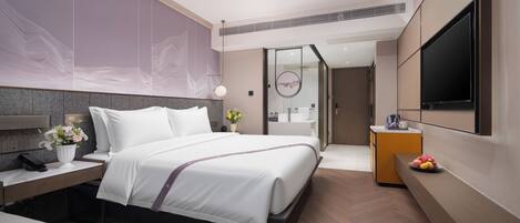 Quarto de hotel moderno com uma cama grande, uma mesinha de cabeceira, televisão fixada na parede e um banheiro visível através de uma porta de vidro.