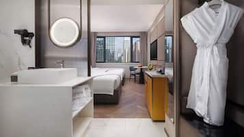 A modern hotel bathroom with a large mirror, a sink, a robe hanging, and a view of the cityscape through the window.