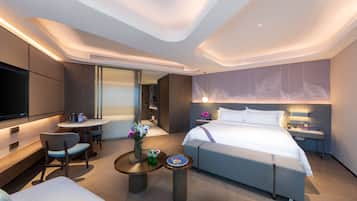 A modern hotel room with a large bed, a desk, a TV, and a seating area with chairs and a table.