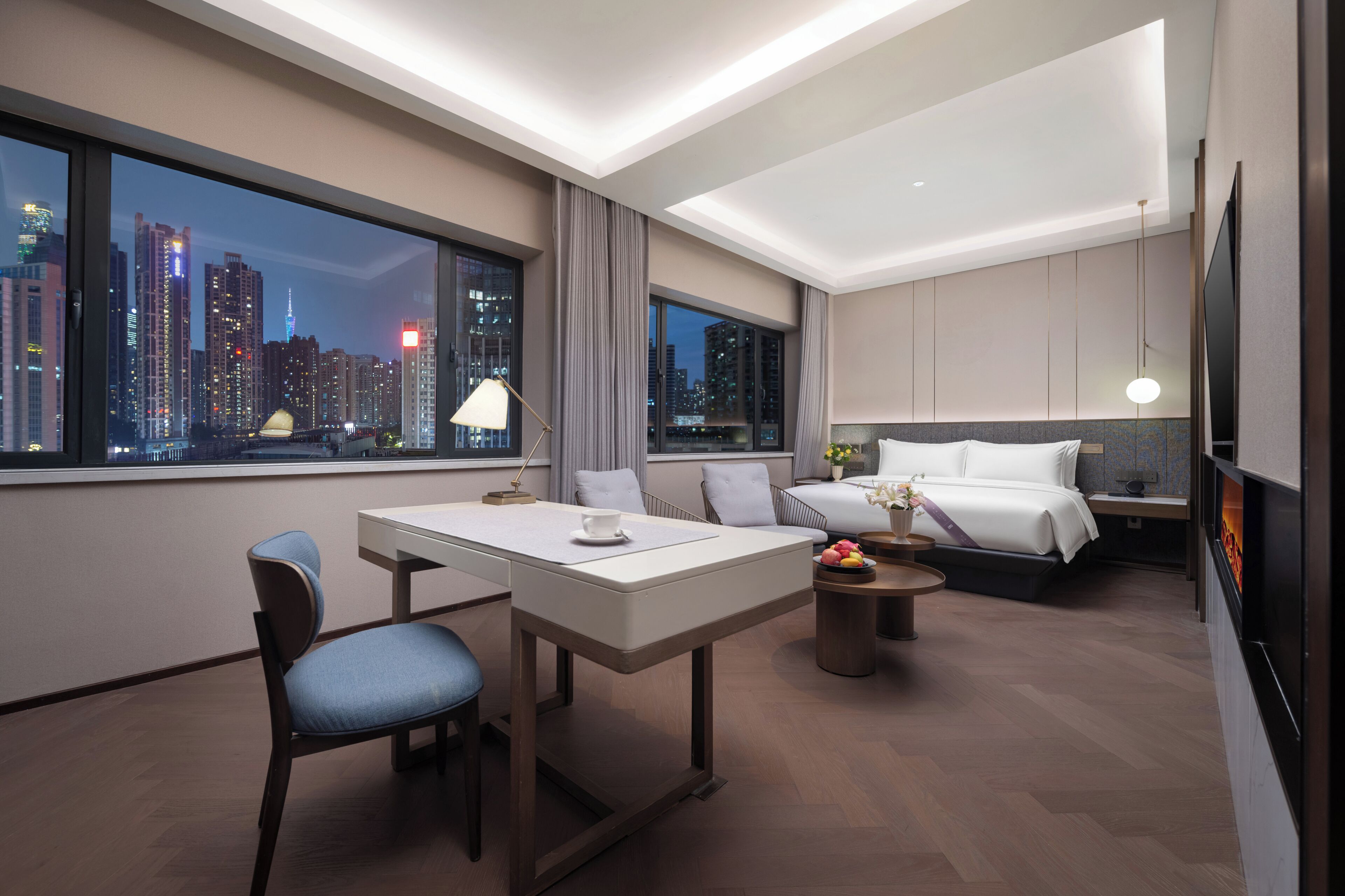 A modern hotel room with a city view, a dining area, and a bed with a nightstand.