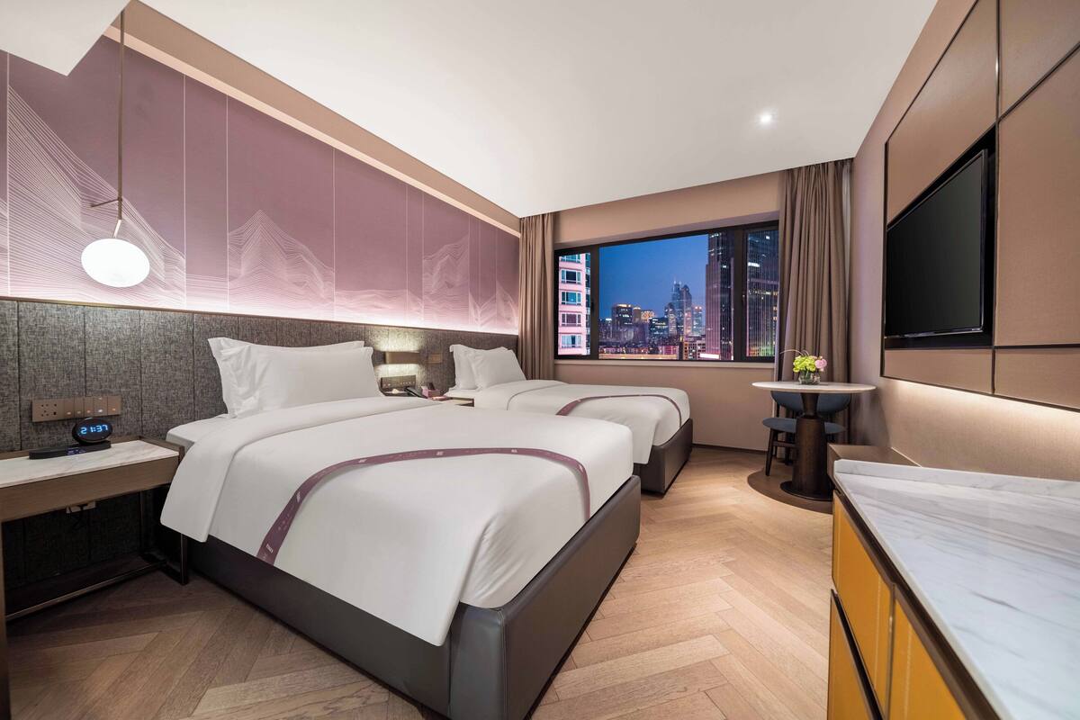 Habitació d'hotel moderna amb dos llits, una finestra gran amb vista a la ciutat, un televisor de pantalla plana i una taula de dinar petita.