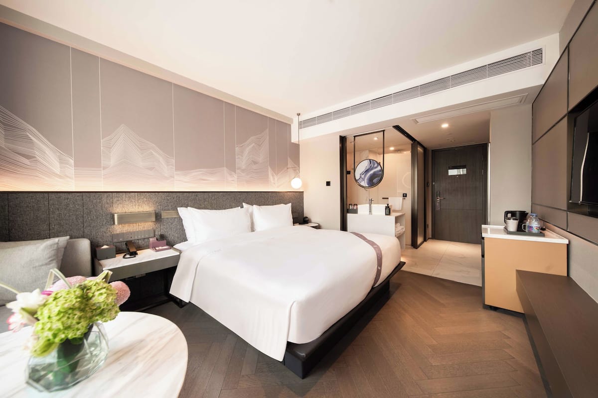 Habitació d'hotel moderna amb un llit gran, una taula de nit amb una làmpada, un escriptori petit i un bany visible a través d'una porta oberta.