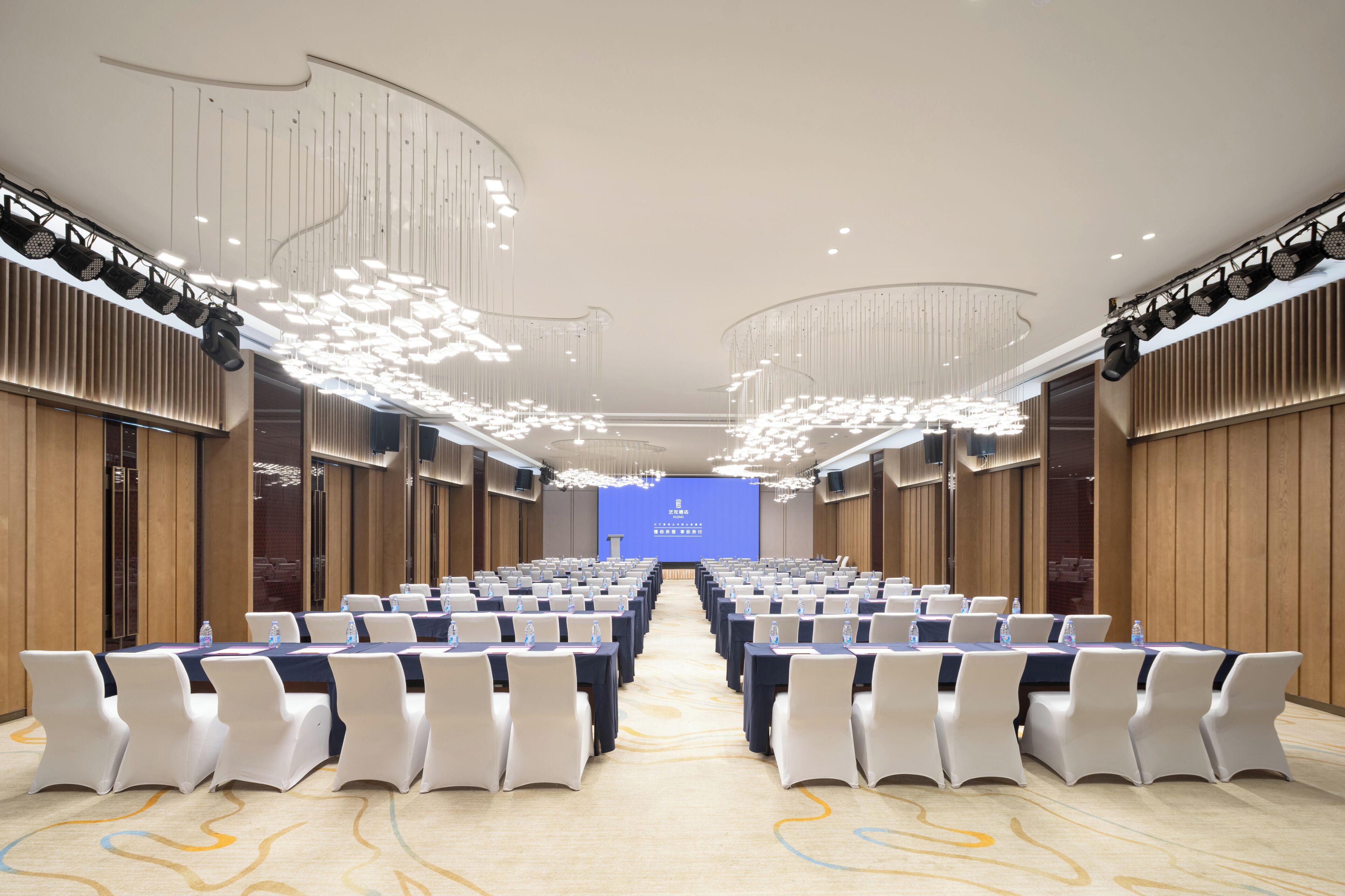 A conference room with rows of white chairs, a blue screen at the front, and modern lighting fixtures.