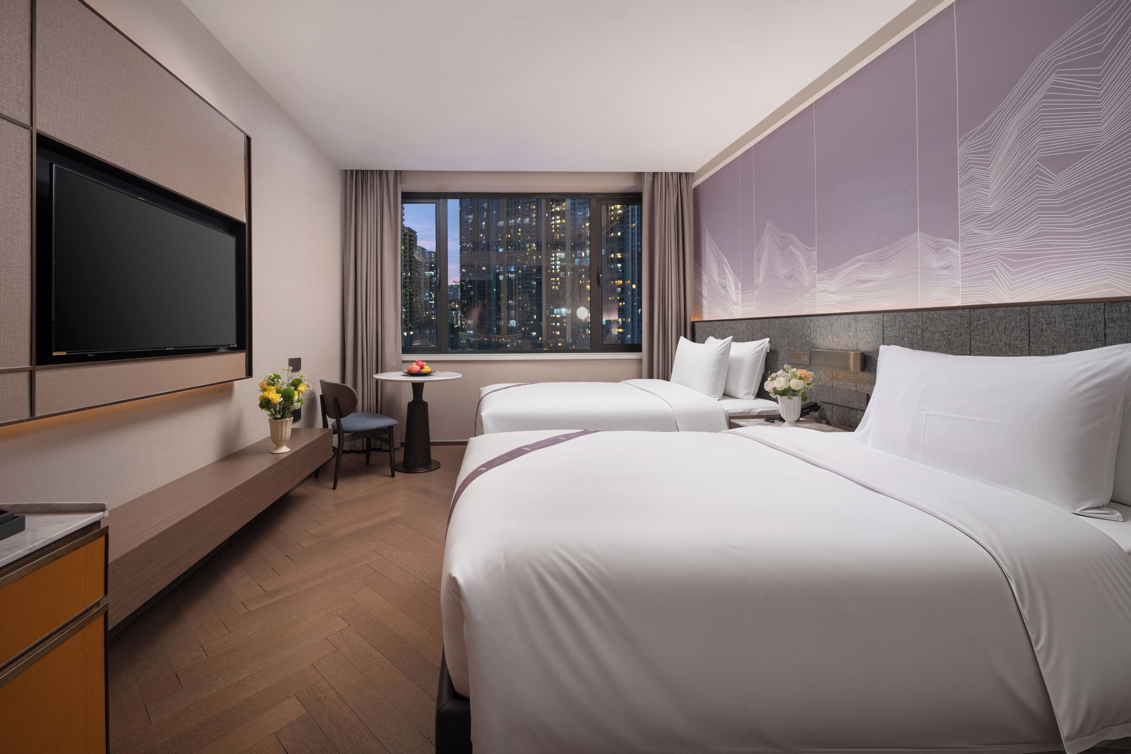 Ein Hotelzimmer mit zwei Betten, einem Schreibtisch, einem Stuhl, einem Fernseher und einem Fenster mit Blick auf die Stadt.