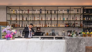 A bar area with a bartender, a marble counter, shelves with liquor bottles, and a flower arrangement.