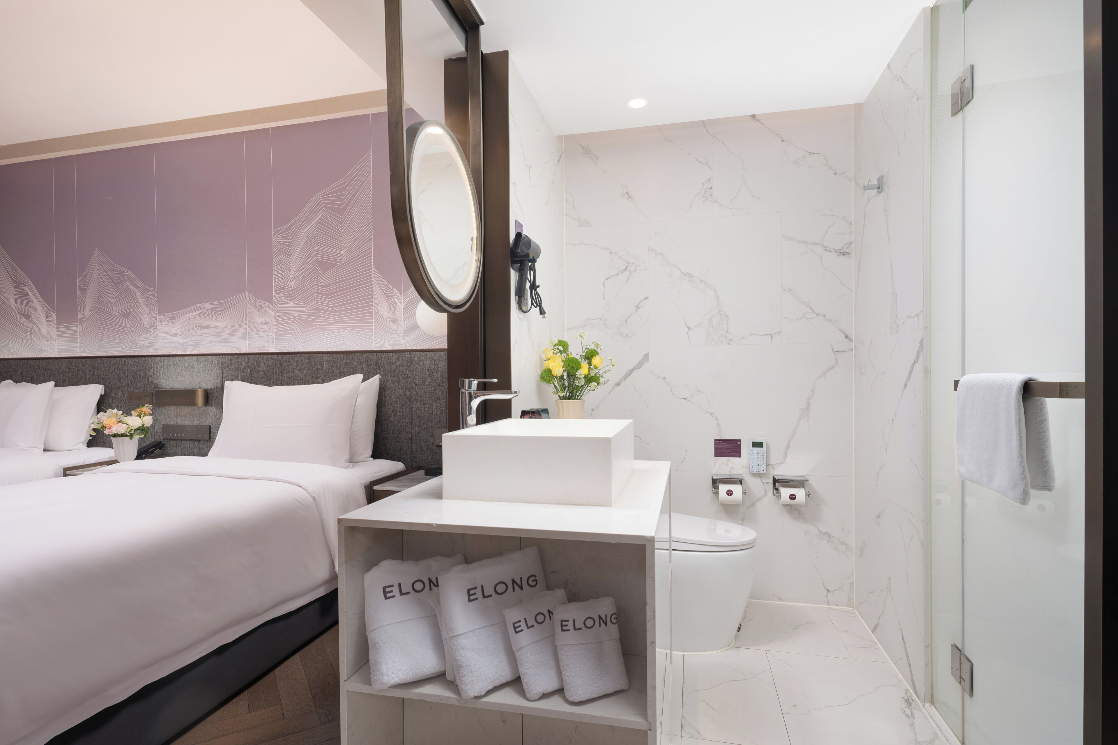 A modern hotel room with a large bed, a bathroom with a marble wall, a round mirror, and a shelf with towels.