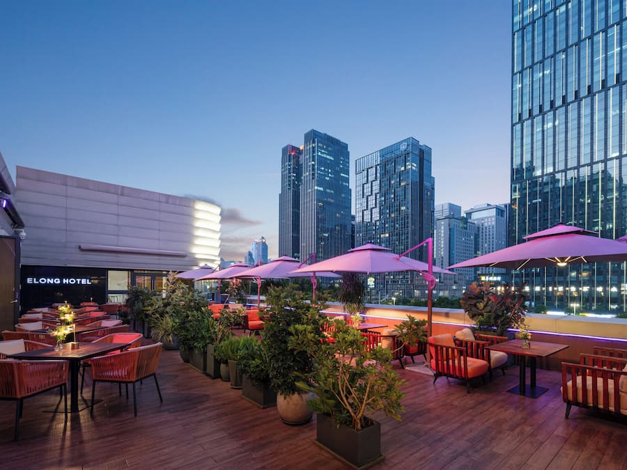 Uma área de estar ao ar livre com mesas e cadeiras, rodeada por plantas em vasos, com vista para um horizonte urbano com edifícios modernos.