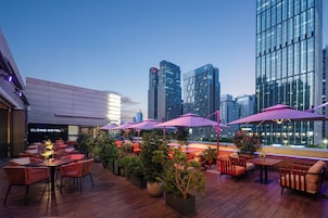 An outdoor seating area with tables and chairs, surrounded by potted plants, and overlooking a cityscape with modern buildings.