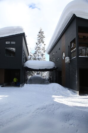 A snow-covered street with two buildings, one with a garage door open. - Alpine Tenshi Villa (Hakuba)