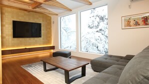 A modern living room with a wooden ceiling, a flat-screen TV mounted on the wall, a grey sofa, a wooden coffee table, and large windows with a view of snow-covered trees. - Alpine Tenshi Villa (Hakuba)
