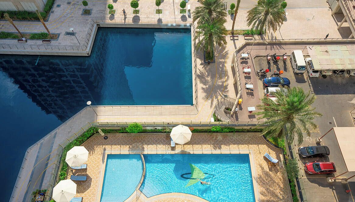 An aerial view of a hotel with a large swimming pool, palm trees, and a parking area.