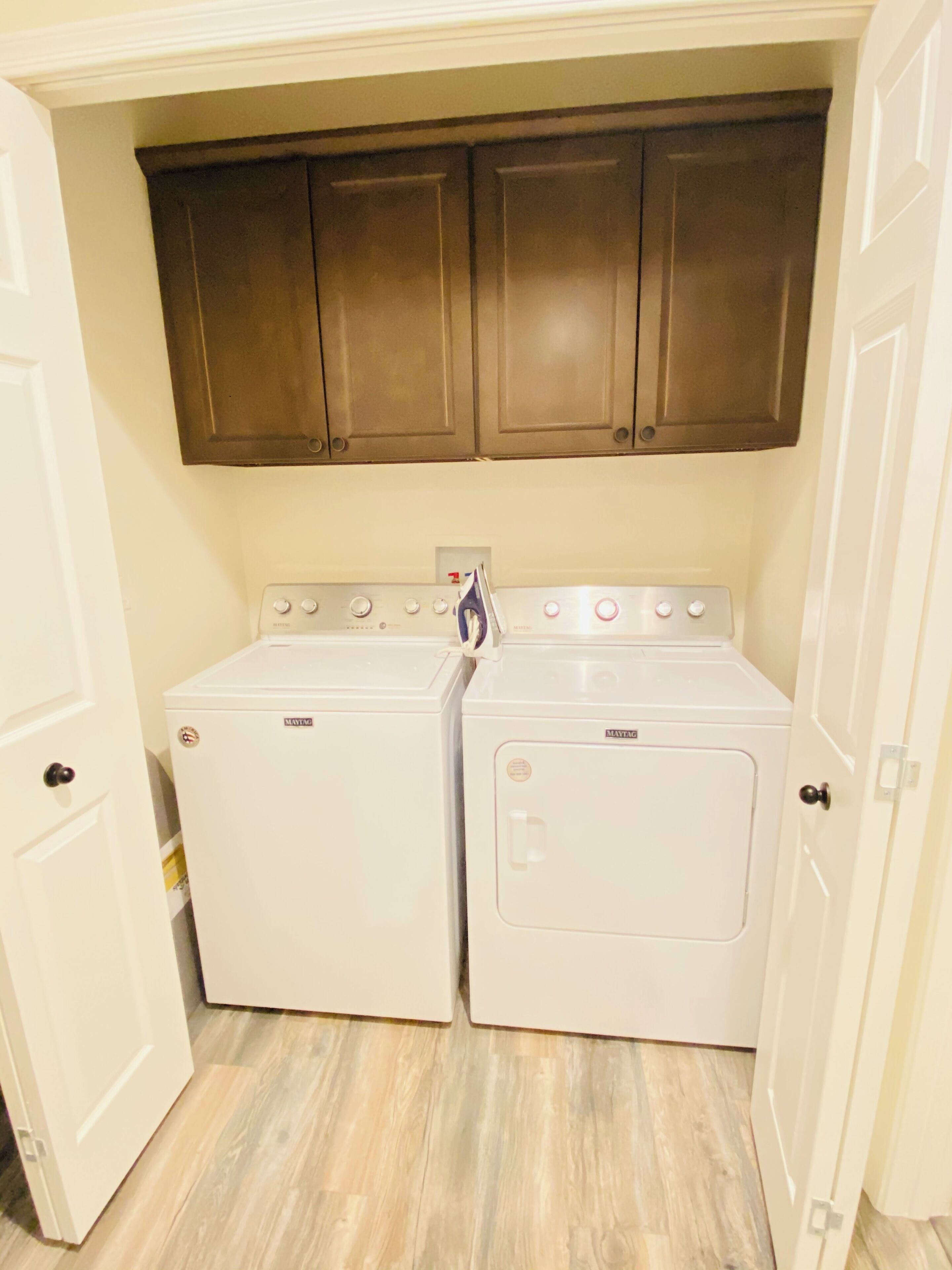 A laundry room with two Maytag washers and dryers, wooden cabinets above, and a light-colored floor.