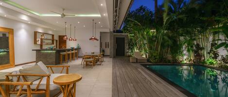 Un área de piscina interior moderna con piso de madera, asientos tipo sala de estar y una zona de bar.