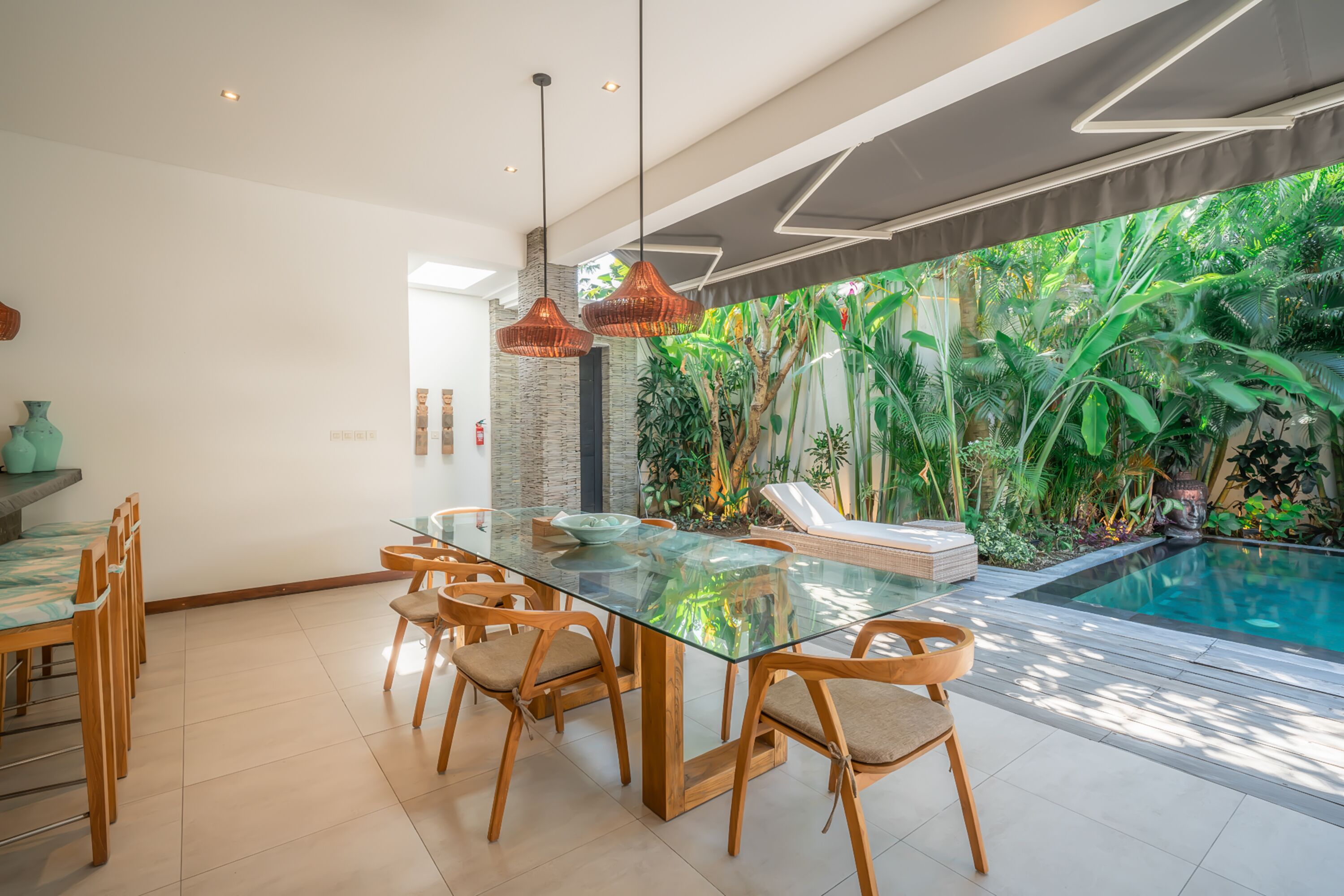 A modern dining area with a glass table and wooden chairs, a poolside lounge, and lush greenery visible through large windows.